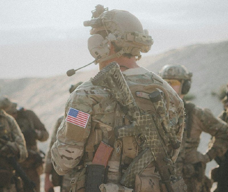 A soldier in camouflage gear, helmet, and headset, with a U.S. flag patch, stands facing a mountainous landscape alongside other uniformed personnel.