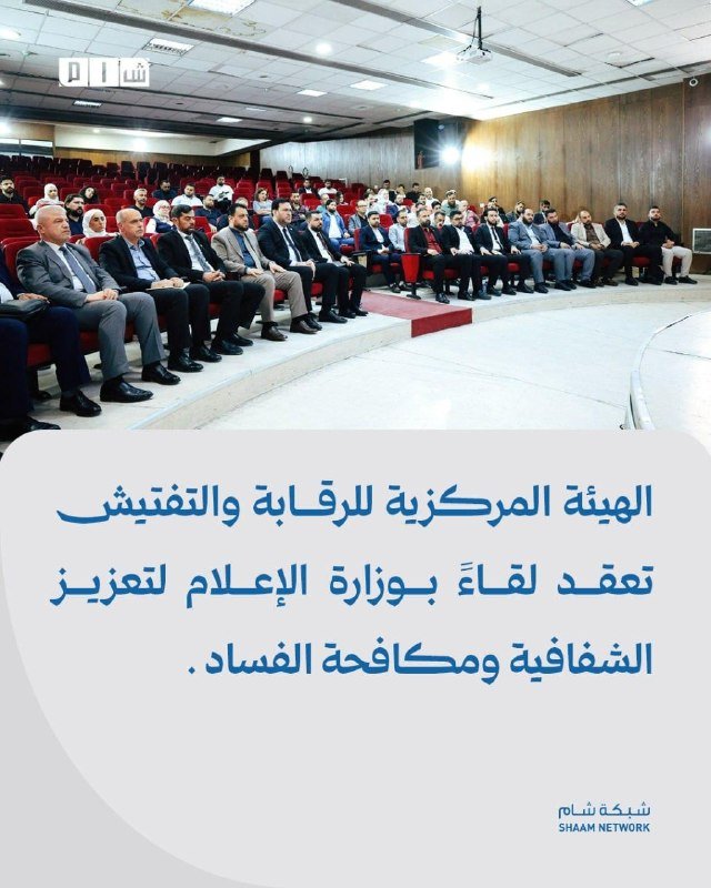 Attendees, including men in suits and a few women, sit in a wood-paneled auditorium facing forward.