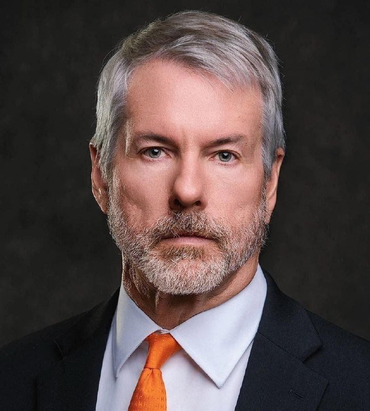 A gray-haired man with a beard, wearing a dark suit, white shirt, and orange tie, poses for a formal portrait against a dark background.