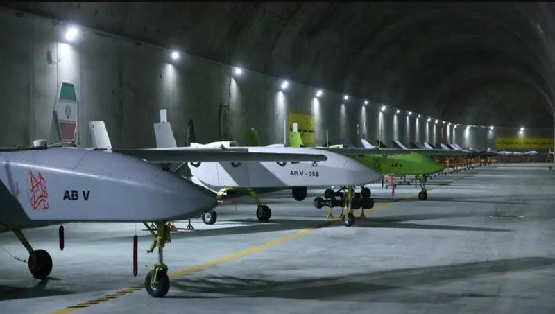 A row of military drones, including one marked "AB V" and another labeled "AB V-005," are parked inside a large illuminated underground hangar.