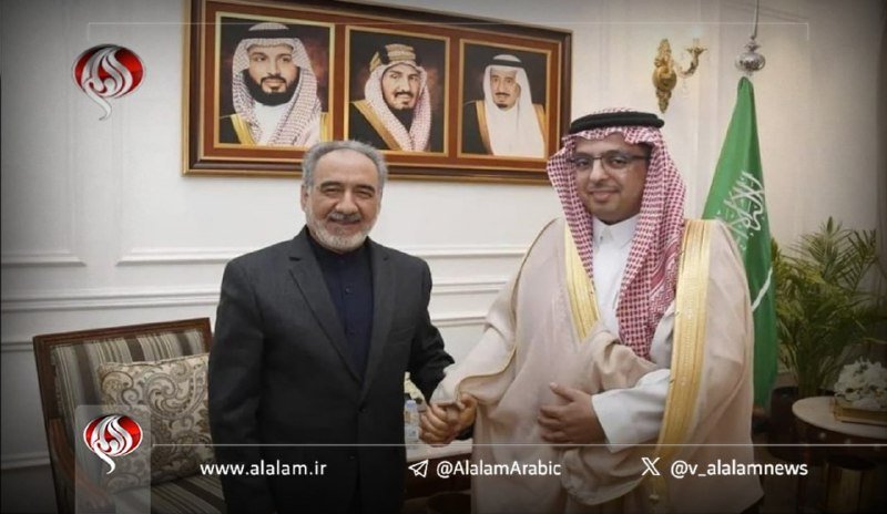 Two men in formal attire shake hands in an ornately decorated room featuring three framed portraits on the wall and a green flag with Arabic script, while network logos appear in the corners.
