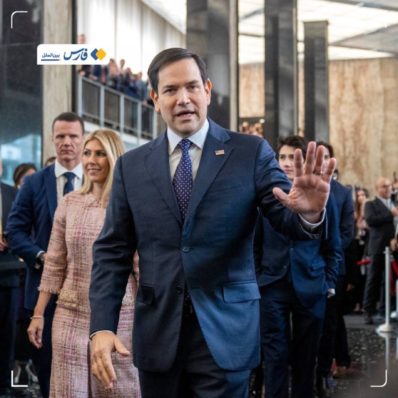 A man in a dark suit and blue tie raises his hand while walking alongside a blonde woman in a pink tweed outfit, followed by a group through a modern building.