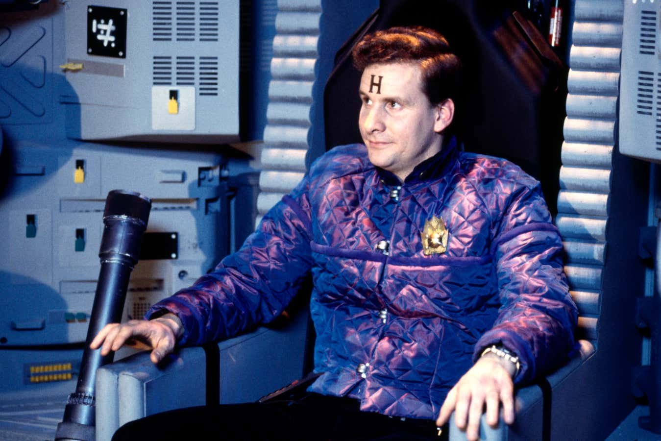 A man with an "H" on his forehead sits in a chair within a metallic, sci-fi style interior, wearing a purple and red patterned jacket with a gold emblem.