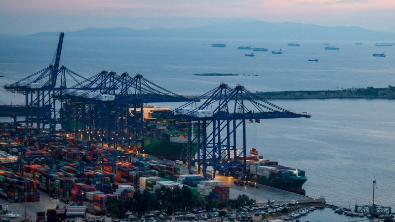 Large cranes load cargo containers onto a green ship docked at a coastal port, with numerous vessels anchored offshore at dusk.