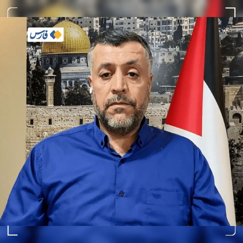 A bearded man in a blue shirt speaks in a studio, with a Palestinian flag beside him and a backdrop showing the Dome of the Rock.