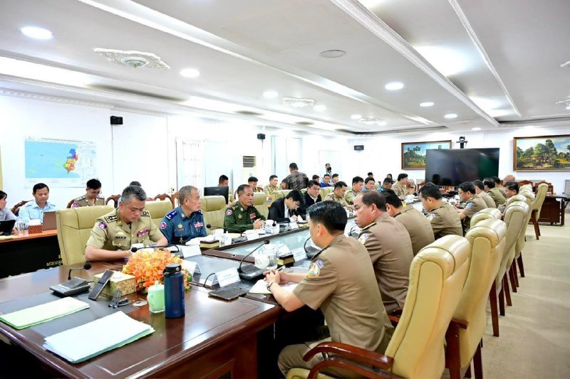 Uniformed officials in tan and green sit around a long conference table with microphones, documents, and water bottles in a brightly lit meeting room.