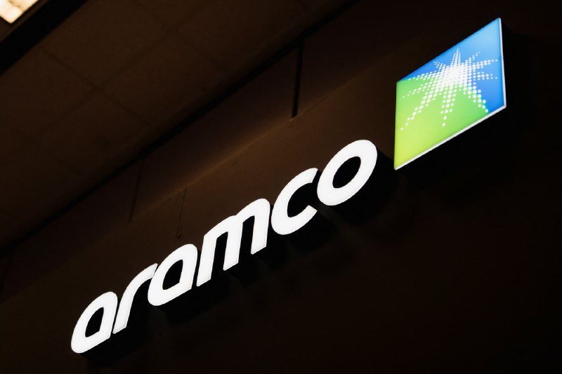 An illuminated "aramco" logo with a green and blue starburst emblem is mounted on a dark exterior wall at night.