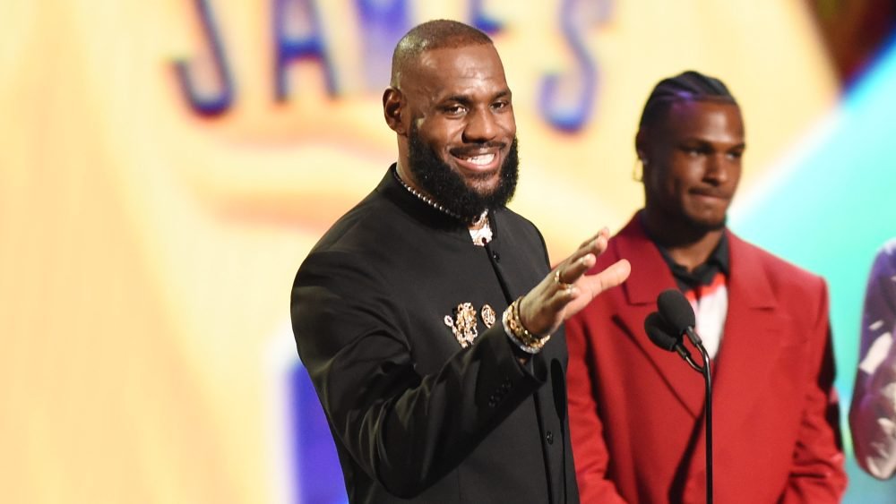 A smiling bearded man in a black suit gestures toward a microphone at a stage event, while another man in a red suit stands behind him.