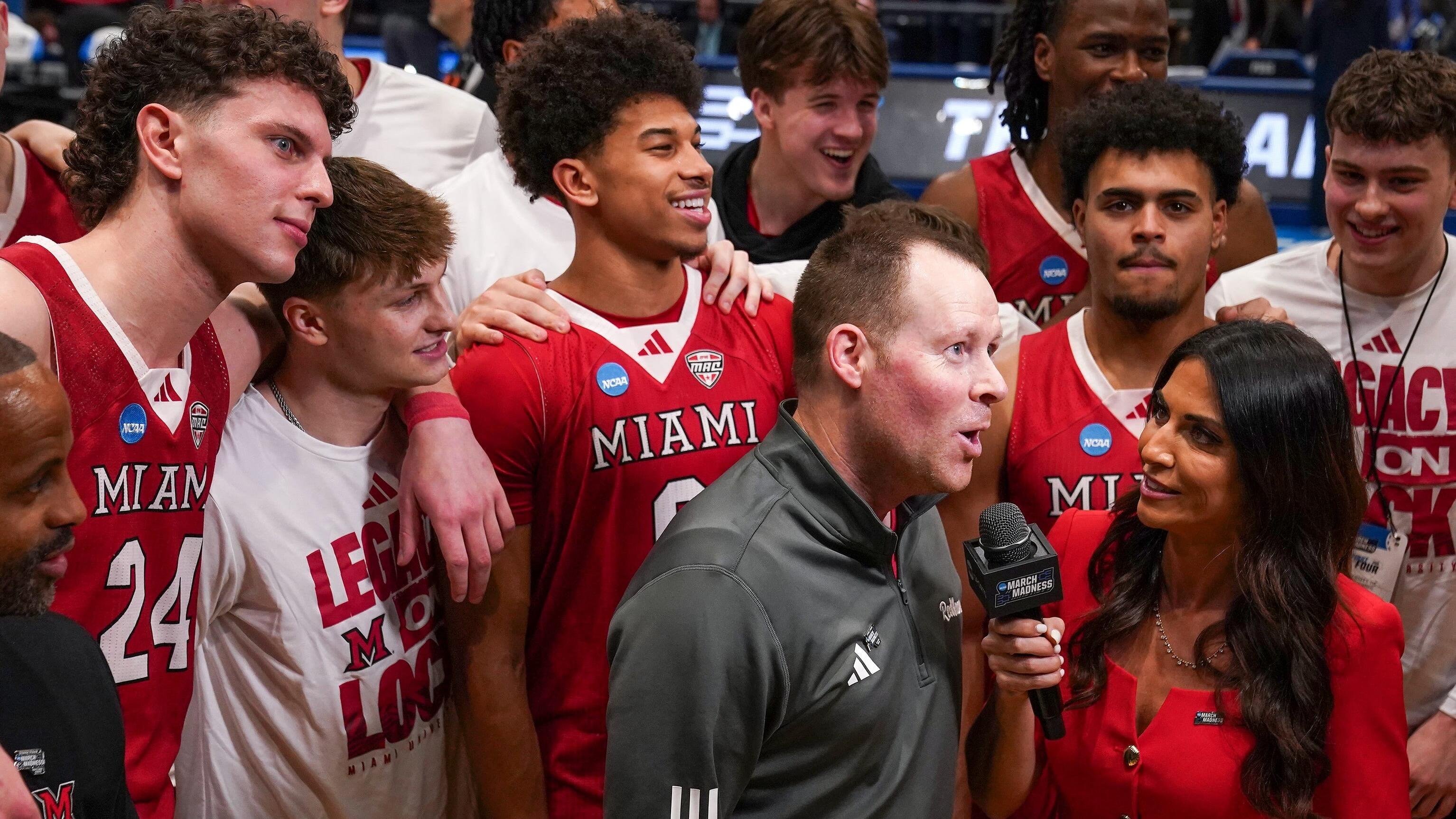 A man in a gray jacket is interviewed by a woman holding a "March Madness" microphone, surrounded by smiling players in red "Miami" jerseys and white "Legacy" shirts.