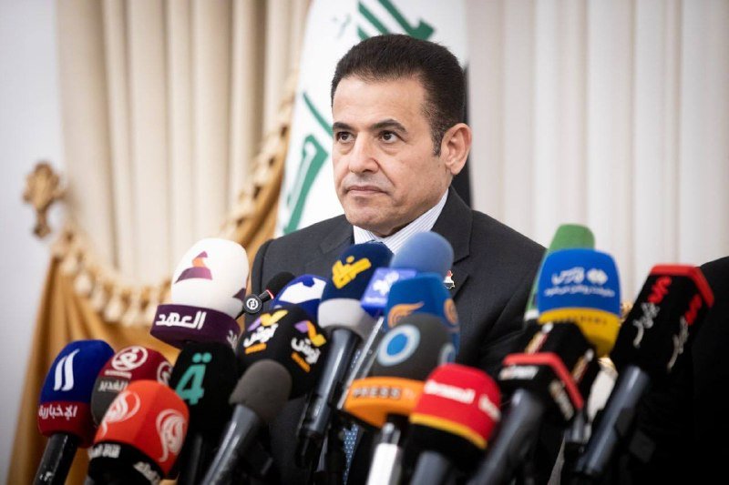 A man in a dark suit speaks at a podium lined with numerous microphones from various media outlets, with an Iraqi flag visible behind him.