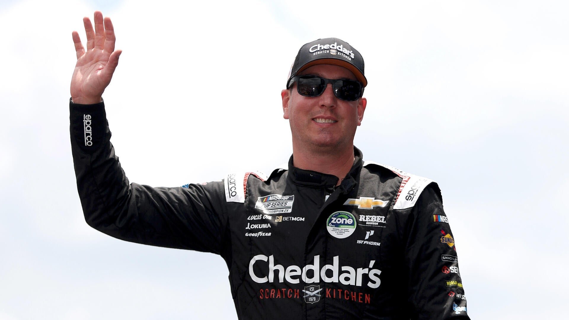 A man in a black Cheddar's Scratch Kitchen NASCAR racing suit, sunglasses, and matching cap waves his hand against a bright sky.