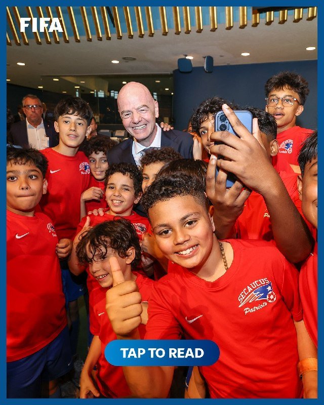 A smiling bald man in a suit poses for a selfie with a group of young soccer players in red jerseys indoors.