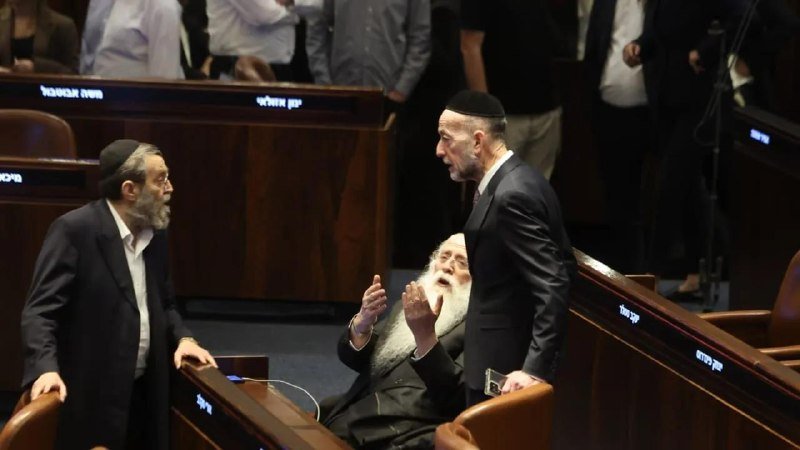 Three men wearing skullcaps converse in a wood-paneled legislative chamber with Hebrew-labeled desks.