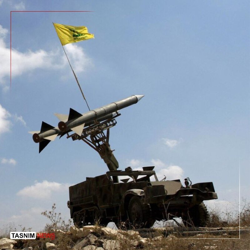 A military truck-mounted missile launcher elevated with two missiles and a yellow flag flies against a partly cloudy sky over a rocky field.