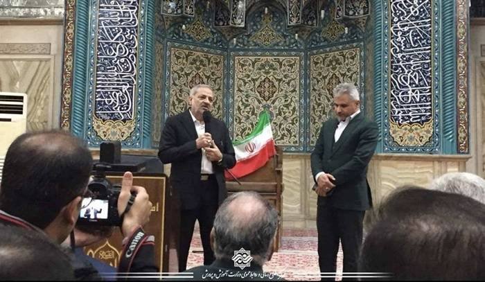 Two men stand before an ornately tiled wall with Arabic calligraphy, one speaking into a microphone beside an Iranian flag, while a photographer and seated audience capture the moment.