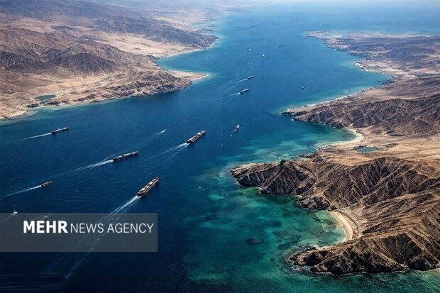 Aerial view of several ships sailing through a narrow turquoise waterway flanked by arid, mountainous coastlines.