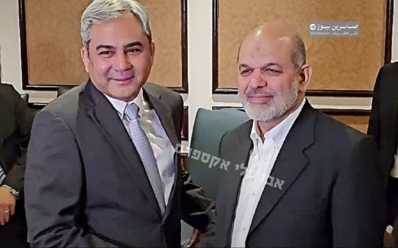 Two men in suits pose together for a photograph indoors, with one wearing a light blue tie and the other in an open-collar white shirt, alongside Hebrew text overlay.