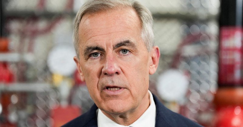 A gray-haired man in a dark suit and white shirt speaks at a podium, with an out-of-focus chain-link fence and orange objects visible behind him.