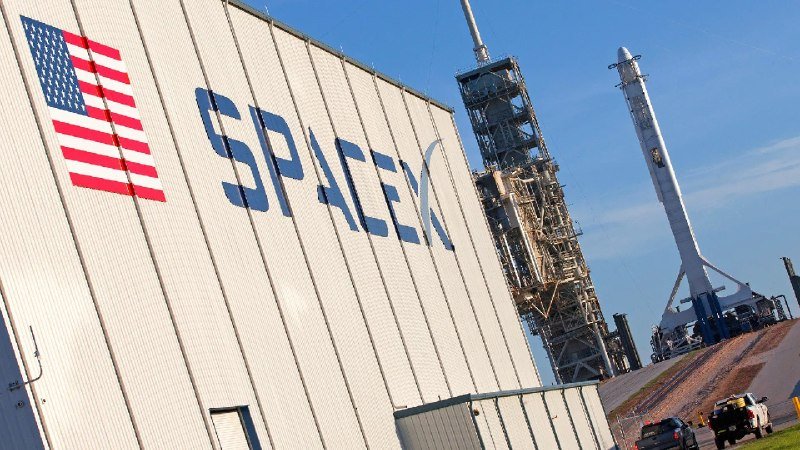 A building displaying a SpaceX logo and American flag stands beside a launch tower and rocket on the pad under a clear blue sky.