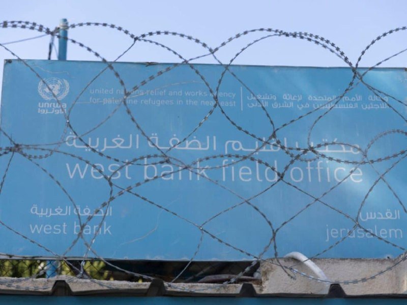 A blue UNRWA sign reading "West Bank Field Office" is visible behind coils of razor wire against a pale sky.