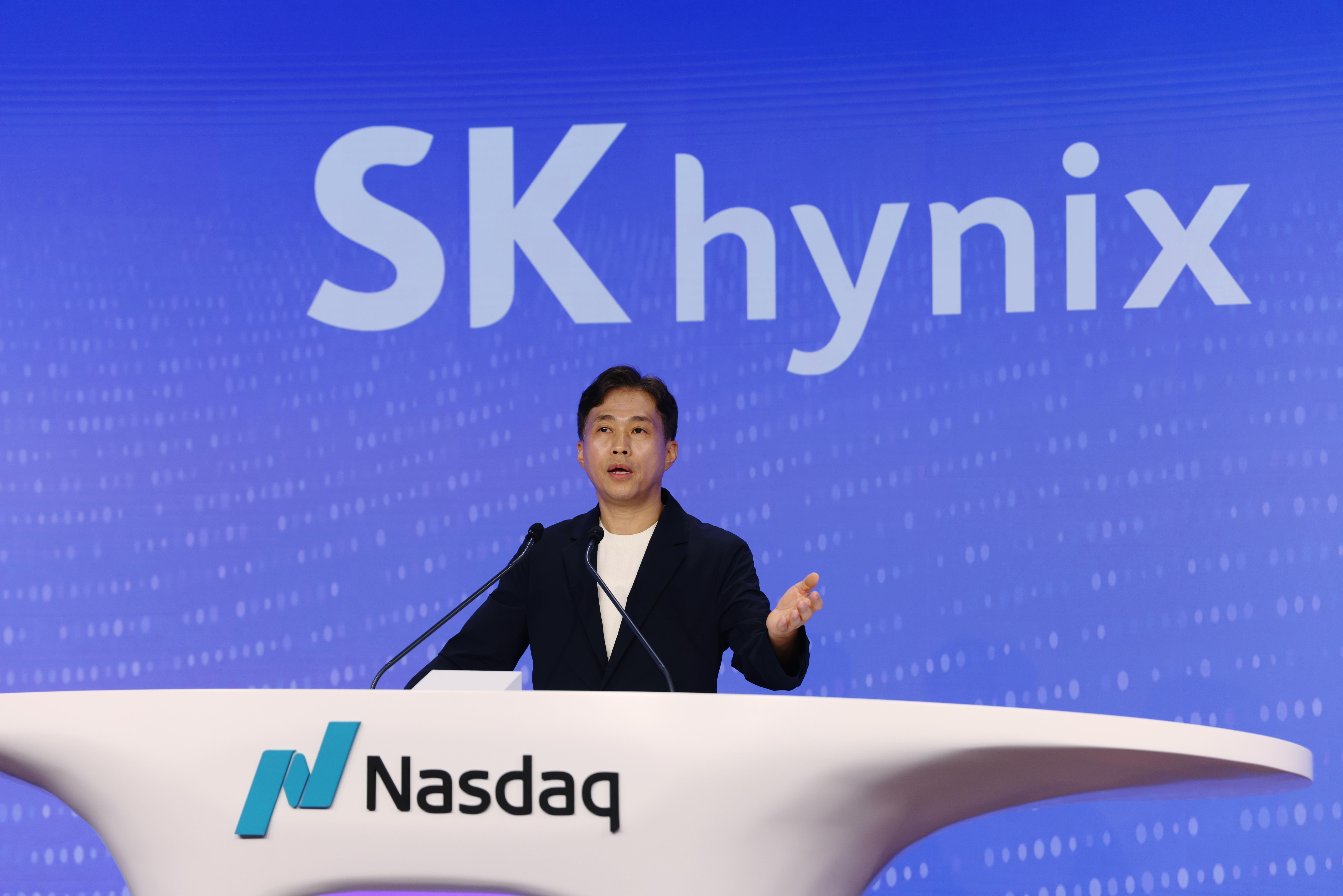 A man in a black blazer speaks at a white podium displaying the Nasdaq logo, with a large "Sk hynix" sign behind him.