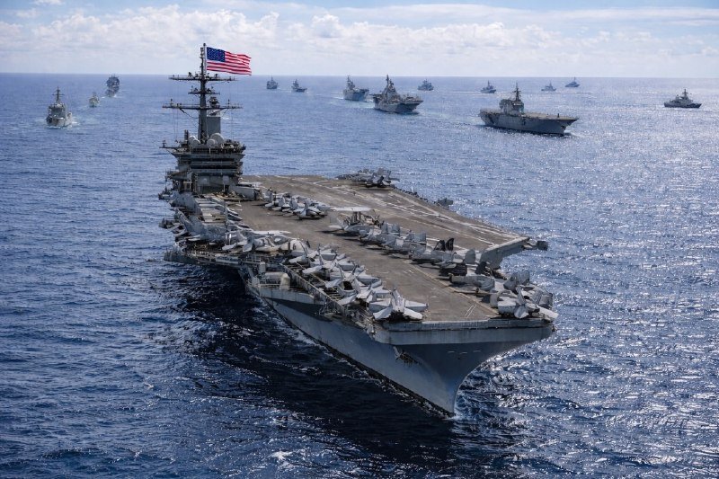 A large aircraft carrier flying an American flag sails through open ocean, accompanied by a fleet of numerous other naval vessels visible in the background.