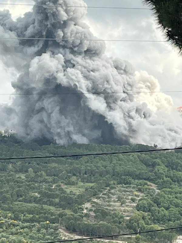 A massive plume of gray smoke billows into the sky above a forested, hilly landscape, with power lines crossing the foreground.