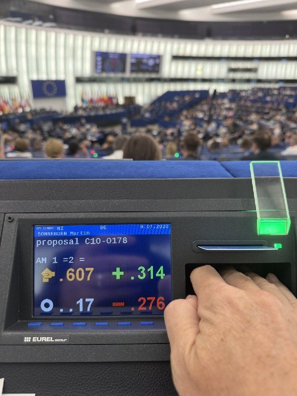A hand inserts a ballot into an electronic voting machine displaying Spanish "proposal" text, inside a large European Parliament chamber filled with attendees.