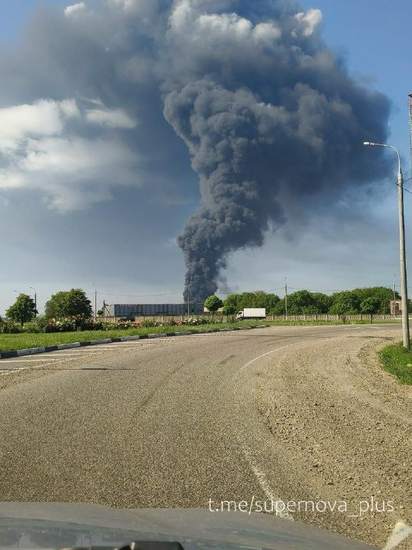 A massive plume of thick black smoke rises into a partly cloudy sky behind a road, trees, and a parked truck.