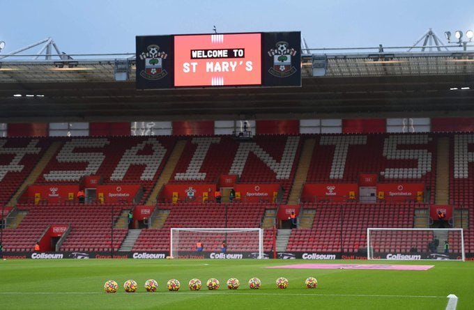 An empty football stadium features a scoreboard reading "WELCOME TO ST MARY'S" with club crests, red seats spelling "SAINTS," and several soccer balls lined up on the pitch near a goal.