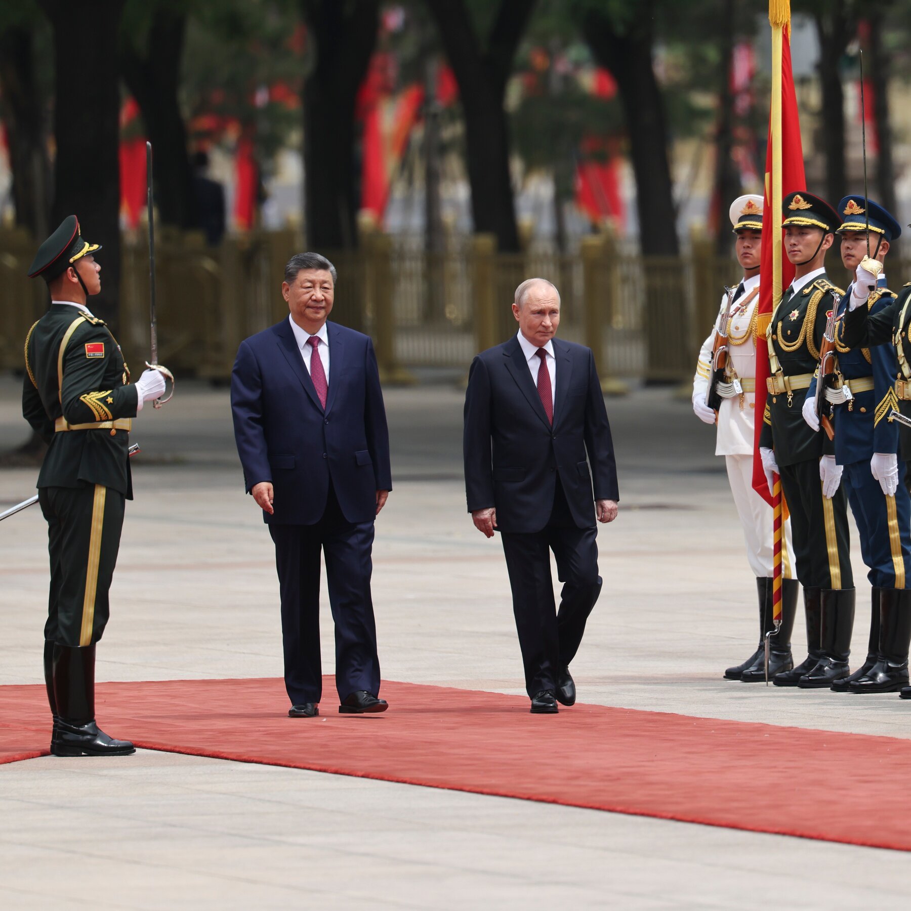 Two men in dark suits walk side-by-side on a red carpet during a military honor guard ceremony, flanked by uniformed soldiers holding swords and flags.