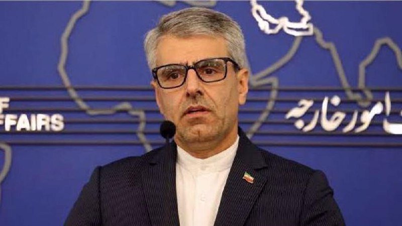 A bespectacled man in a dark suit with a flag lapel pin speaks at a podium before a blue backdrop featuring a map outline and partial text reading "AFFAIRS."