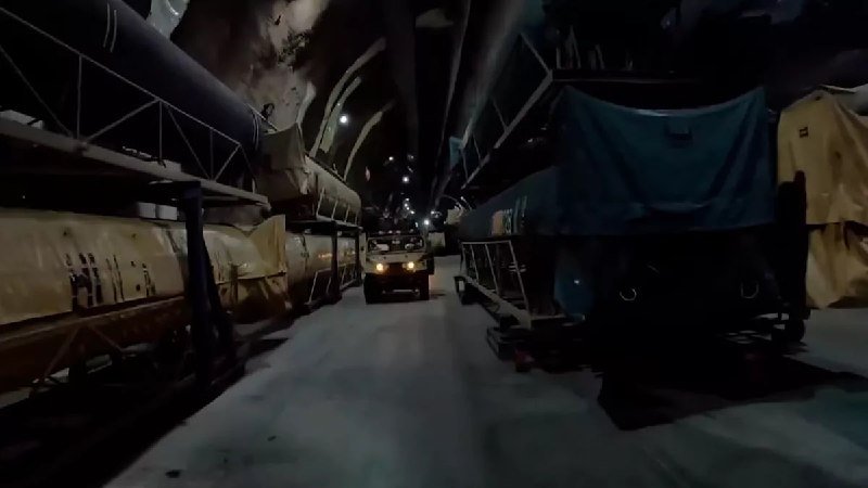 A white truck with headlights on navigates a dimly lit, arched tunnel between rows of parked heavy machinery and equipment.