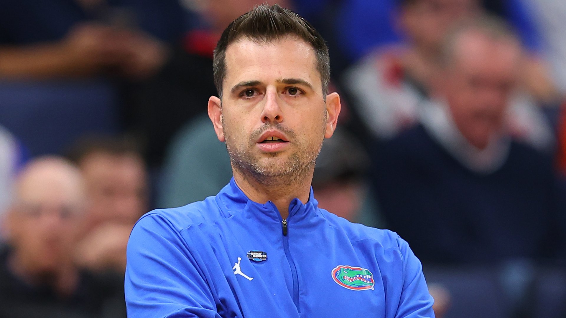 A man in a blue Florida Gators quarter-zip jacket with a Jordan logo looks on against a blurred crowd background.