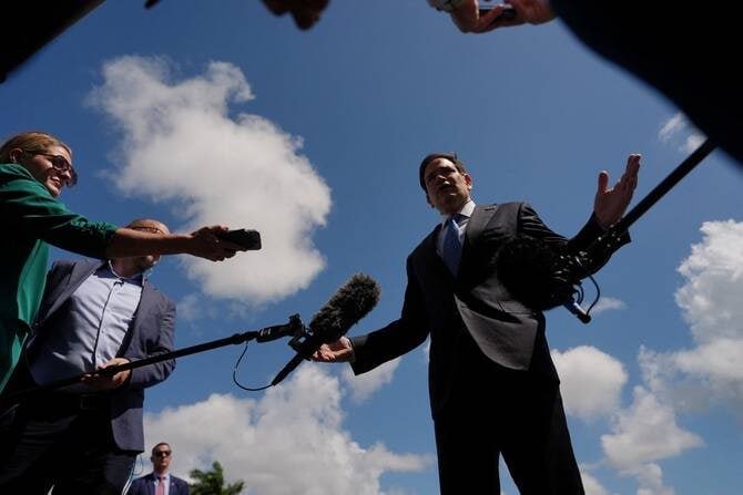 A man in a dark suit and blue tie speaks to reporters holding microphones and a camera against a cloudy blue sky.
