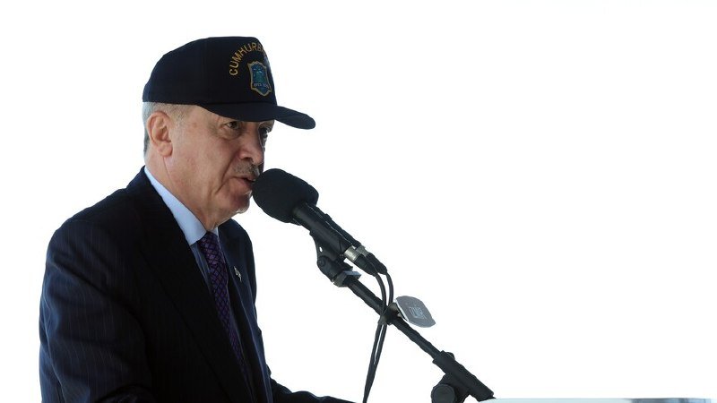 A man wearing a dark cap and suit speaks into a microphone against a white background.