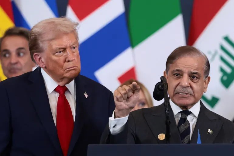 Two men in dark suits stand before international flags, one at a microphone gesturing while speaking, the other beside him looking on.
