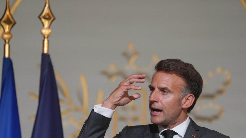 I cannot identify the person in the image. I can describe what I see: a man in a dark suit and white shirt gestures with his raised right hand, standing before two blue flags with gold spear-tipped finials and a pale wall with gold floral motifs.