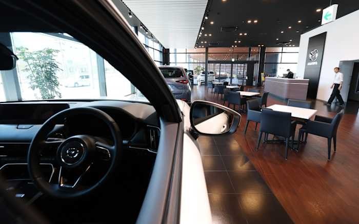 The interior of a vehicle with a steering wheel is visible in the foreground, overlooking a car dealership showroom with seating areas, other vehicles, and a man walking.