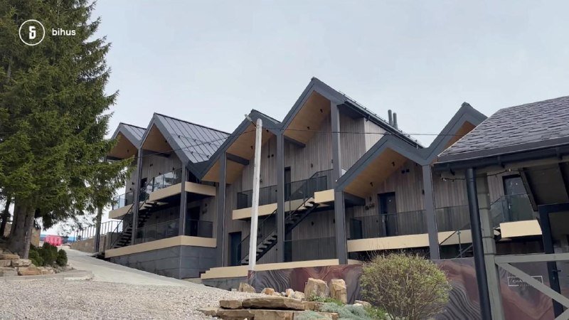 A row of modern multi-story townhouses with steep gabled roofs, dark wood siding, and glass-railing balconies sits along a paved road beside a tall evergreen tree.