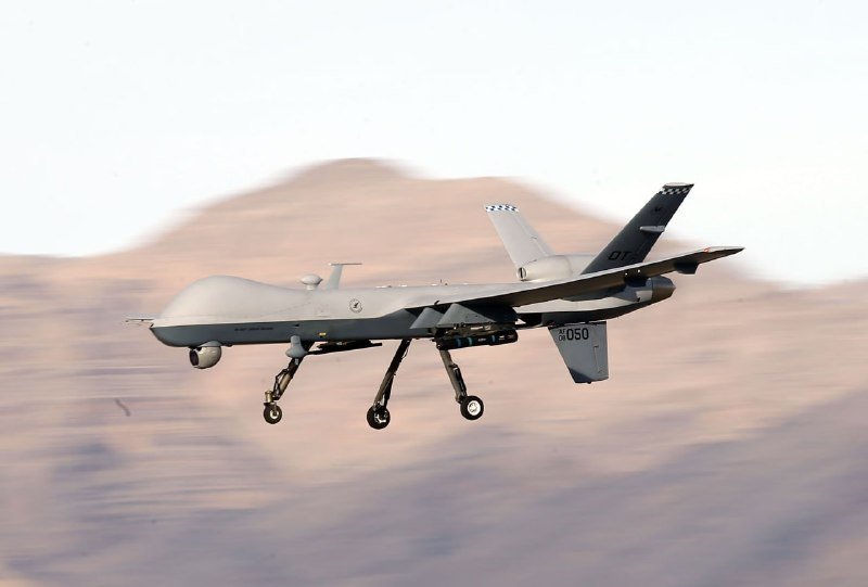 A gray military drone marked "050" flies low with landing gear deployed over a mountainous desert landscape.