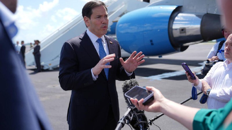 A man in a dark suit and blue tie gestures with both hands while speaking to reporters holding smartphones, standing on a tarmac beside a blue and white aircraft with boarding stairs.
