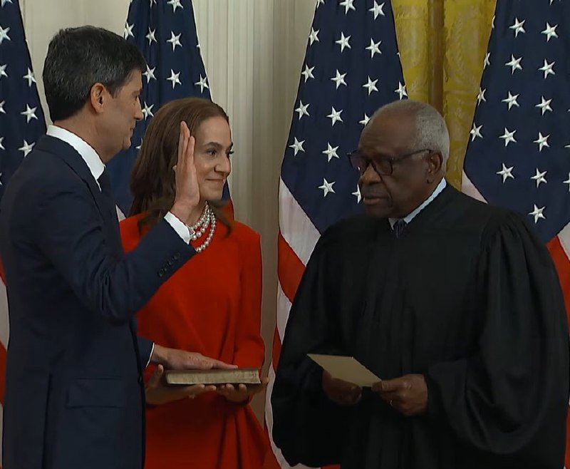 A man in a dark suit raises his right hand while a woman in a red dress holds a book, facing an older man in a black judicial robe holding papers, with American flags in the background.