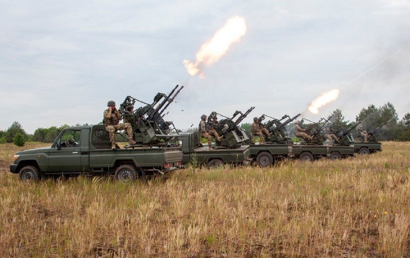 A line of green pickup trucks mounted with anti-aircraft guns fires into the sky, with soldiers in camouflage operating the weapons in a grassy field.