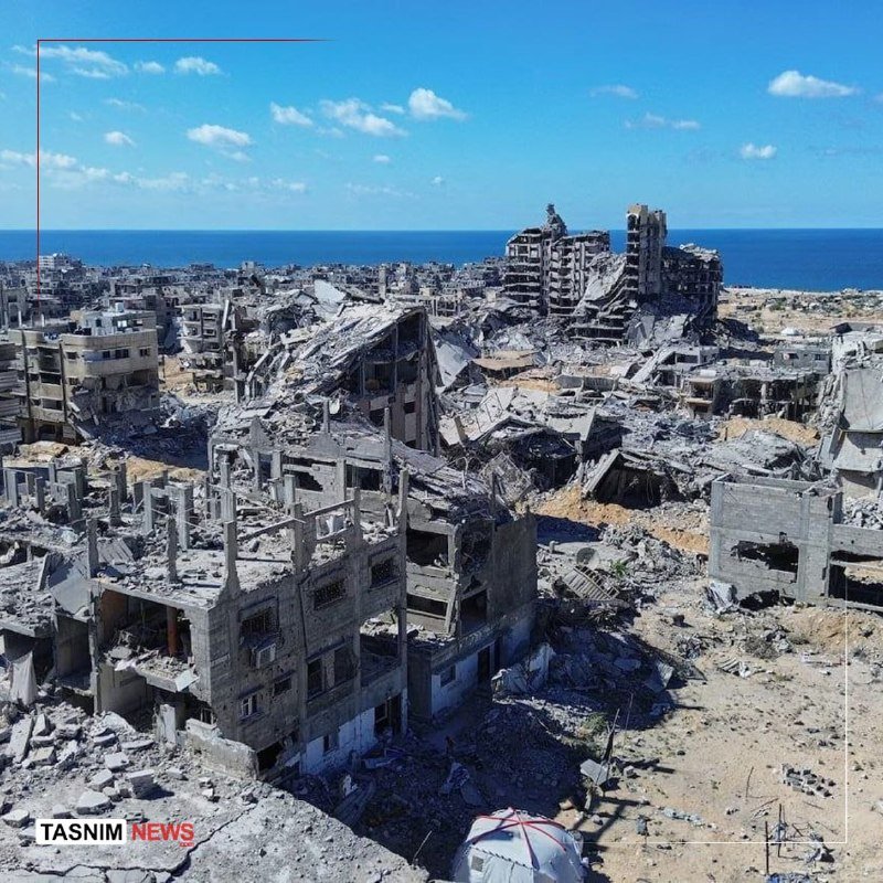 Extensive urban destruction with collapsed and heavily damaged multi-story buildings stretches toward a coastline under a blue sky, with a "TASNIM NEWS" logo visible.