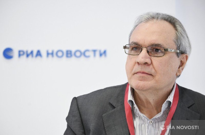 A gray-haired man with glasses, wearing a dark blazer and striped shirt with a red lanyard, looks off-camera in front of a backdrop reading "РИА НОВОСТИ."