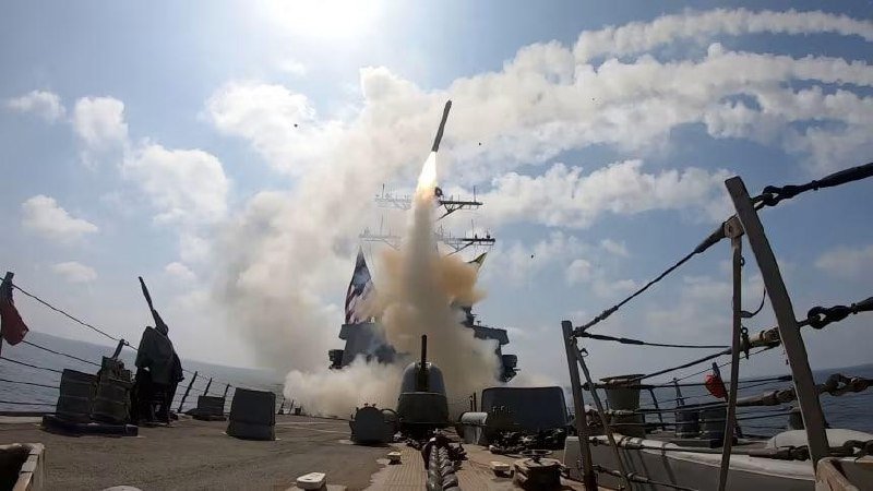 A missile launches vertically from a naval warship at sea, leaving a trail of smoke and flames as crew members watch from the deck.