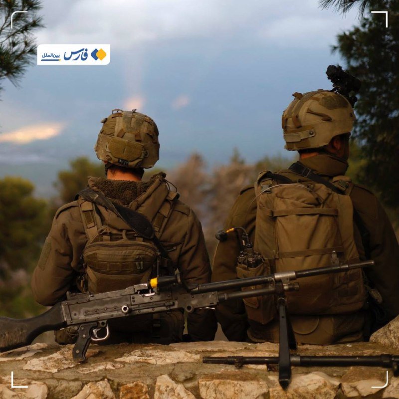 Two soldiers in combat gear and helmets, one carrying a machine gun, sit on a rocky outcrop overlooking a forested landscape at dusk.