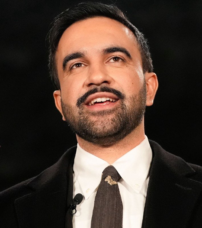 A man with dark hair and a beard, wearing a dark suit, white shirt, and brown tie with a lapel microphone, appears to be speaking against a black background.
