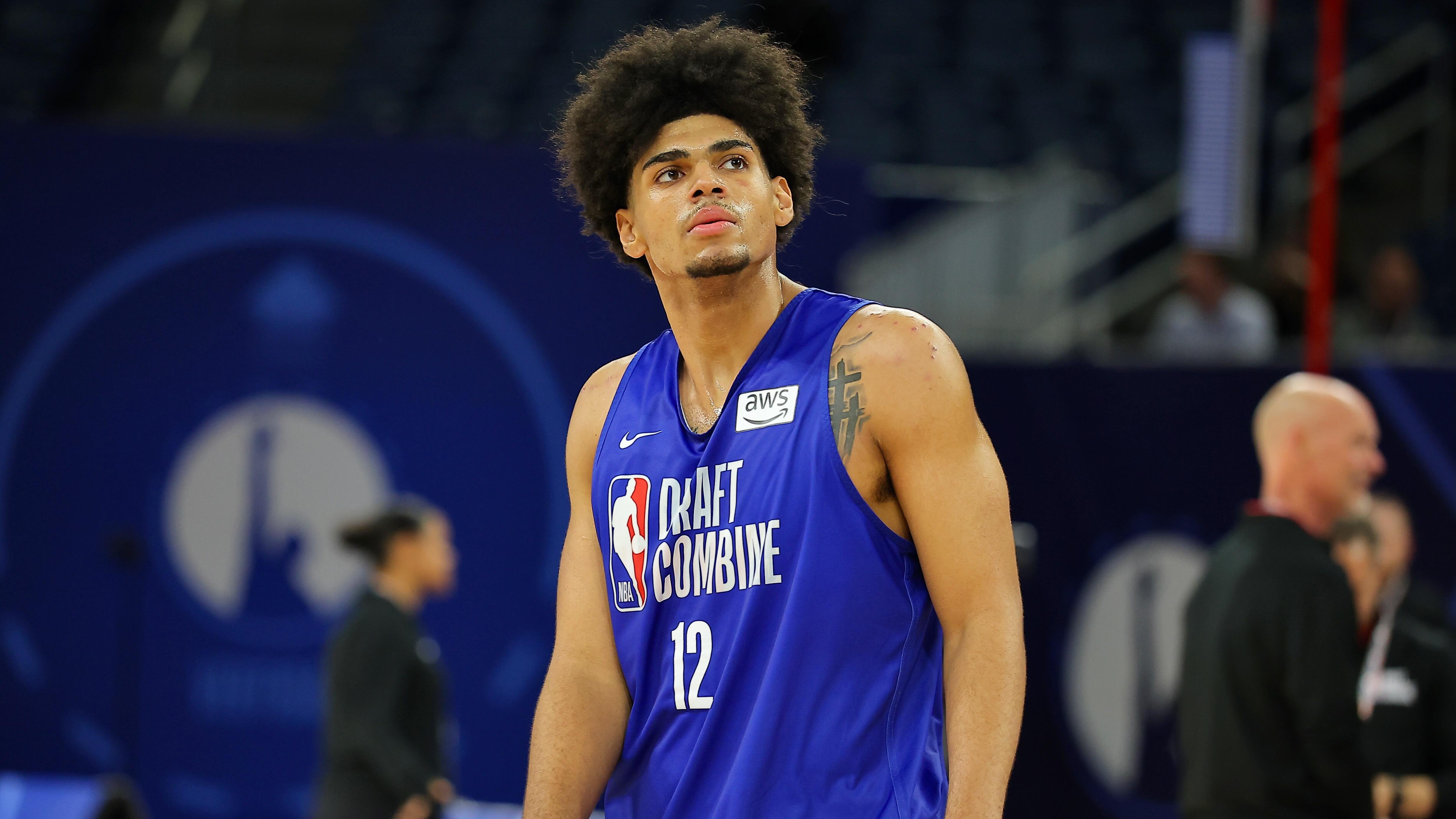 A young basketball player wearing a blue "Draft Combine" jersey with the number 12 stands on a court, looking upward with a focused expression.